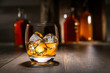 Close up of single malt scotch whiskey, craft bourbon in round tumbler glass, on the rocks, with liquor bottles and rustic background