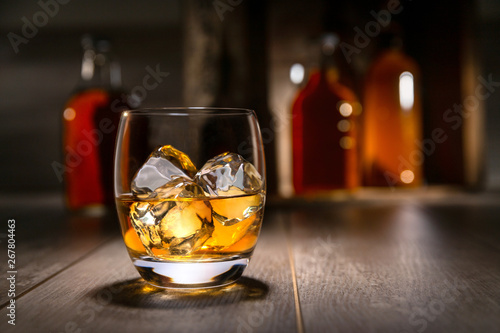 Close up of single malt scotch whiskey, craft bourbon in round tumbler glass, on the rocks, with liquor bottles and rustic background