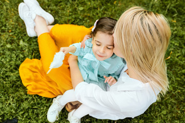 Outdoors image of beautiful mother kissing and cuddling with her daughter, enjoying the time together. Cute little girl embraces her mom, sitting on the green grass in the park. Mothers day