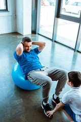 Canvas Print - son helping father sitting on fitness ball and doing sit up exercise at gym