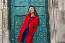 Young Lady In A Red Coat And Sitting In A Denim Suit Holding A Suitcase Luggage, A Trip To Europe In The Center Of The Old Town New Exciting Impressions