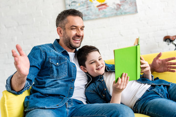 Wall Mural - smiling father and son in denim reading book on couch at home