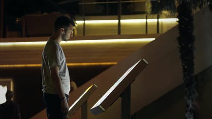 Attractive man using interactive touchscreen terminal display for searching info. Modern teen in wireless earphones. Evening time, lowlight exterior. Education and entertainment concept. Close up shot