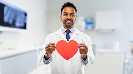 medicine, dentistry and healthcare concept - smiling indian male dentist in white coat with red heart shape and stethoscope over dental clinic office background