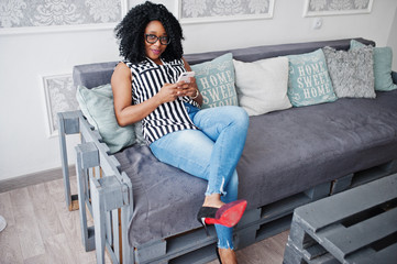 Beautiful african american woman with curly afro hair and eyeglasses, sitting on couch with mobile phone in hand.