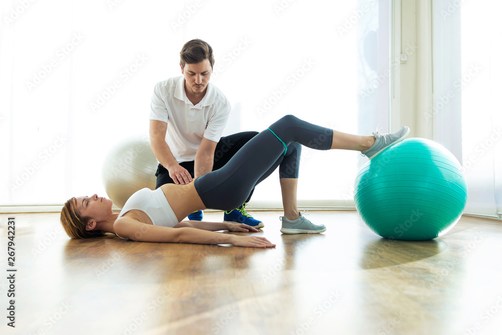 Physiotherapist helping patient to do exercise on fitness ball in physio room. - obrazy, fototapety, plakaty 
