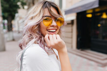 Close-up Portrait Of Attractive Joyful Girl With Amazing Smile Wears Stylish Yellow Sunglasses And White Shirt. Stunning Curly Young Lady With Blonde Hair Excitedly Posing On The Street.