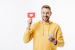 © LIGHTFIELD STUDIOS - cheerful young man in yellow hoodie holding red paper cut card with heart symbol while using smartphone isolated on white