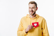 © LIGHTFIELD STUDIOS - handsome cheerful man in yellow hoodie holding red paper cut card with heart symbol isolated on white