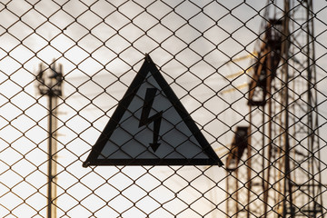 Caution high voltage. A triangular sign with a lightning bolt hanging on a lattice fence. The tower and high-voltage towers are blurred in the background. Evening or morning sky. Silhouette.