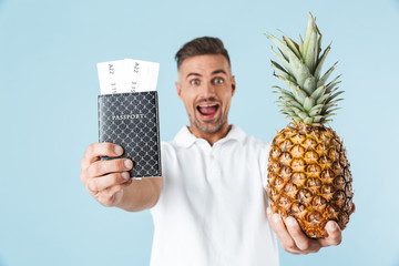 Poster - Handsome excited happy adult man posing isolated over blue wall background holding passport and pineapple.