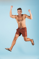 Poster - Excited happy adult man in swimwear posing isolated over blue wall background jumping make winner gesture.