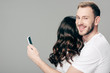 © LIGHTFIELD STUDIOS - handsome smiling man embracing woman and using smartphone isolated on grey