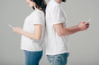 © LIGHTFIELD STUDIOS - cropped view of cheerful man and woman in white t-shirts standing back to back and using smartphones isolated on grey