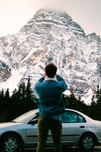 Man Standing Near Gray Sedan Taking Picture Of Snow-covered Mountain