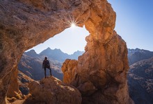Person Standing On Rock Under Arch Formation Rock While Facing Sun