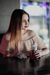 © spaskov - Young beautiful brown-haired girl in a nightclub with neon lights sits by the glass and drinks a red cocktail.