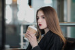 © spaskov - A young beautiful brunette girl in a gray coat is sitting on a bench waiting. Drinking coffee and holding a paper cup with a drink.