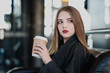 © spaskov - A young beautiful brunette girl in a gray coat is sitting on a bench waiting. Drinking coffee and holding a paper cup with a drink.