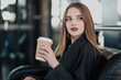 © spaskov - A young beautiful brunette girl in a gray coat is sitting on a bench waiting. Drinking coffee and holding a paper cup with a drink.