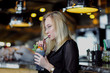 © spaskov - A young beautiful blond woman with glasses sits at the bar in a yellow interior. Drinks alcoholic cocktail with a straw and green mint.