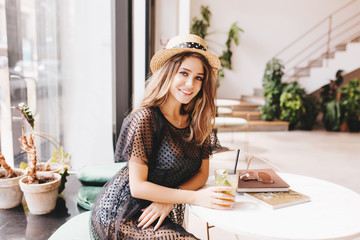 Wall Mural - Graceful young lady resting at the table with glass of cold tea and magazines. Indoor portrait of smiling girl in summer hat posing with cocktail with plants on background.