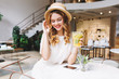 © Look! - Romantic girl with blonde curls resting in cafe and shy smiling, looking down. Indoor portrait of cute young lady in vintage hat holding glass of fruit beverage and sit at the table with phone on it.