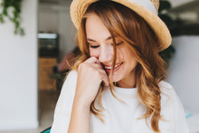 Close-up photo of amazing happy girl with pale skin shy laughing and cover face with hand. Indoor portrait of ecstatic blonde young woman in hat smiling with eyes closed.