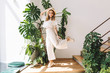 © Look! - Ecstatic blonde girl in white pants funny dancing at home posing in front of big plant. Indoor portrait of jumping young woman in hat and glasses happy smiling next to stairs.