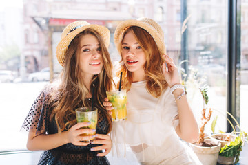 Wall Mural - Close-up portrait of tired but happy girls enjoying cocktails after shopping in weekend. Stunning ladies in vintage attires relaxing together in cafe and drinking juice.