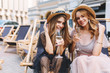 Lightly tanned girls with elegant curly hairstyle drink champagne and smiling. Refined blonde lady in silver bracelet spending time with best friend and resting outdoor on summer chairs.