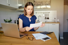 Thoughtful Young Woman Using A Laptop Computer Sitting At Her Kitchen Holding Utility Bill And Bank Statements. Home Interior.