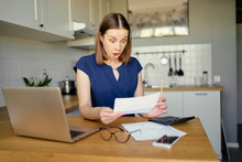 Stressed Over Bills. Portrait Of Surprised Young Woman Using A Laptop Computer Sitting At Her Kitchen Holding Utility Bill And Bank Statements. Home Interior.