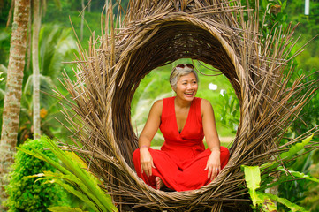 Wall Mural - attractive and happy middle aged 40s or 50s Asian tourist woman with grey hair and elegant red dress sitting outdoors at tropical jungle relaxed admiring beauty of nature