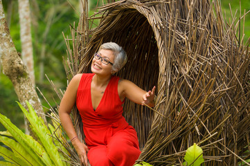 Wall Mural - attractive and happy middle aged 40s or 50s Asian tourist woman with grey hair and elegant red dress sitting outdoors at tropical jungle relaxed admiring beauty of nature