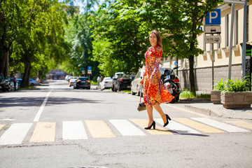 Wall Mural - Young beautiful blonde woman in a red flower dress crosses the road at a crosswalk