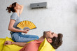 young woman with hand fan sitting on yellow sofa under air conditioner near lying man