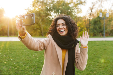 Wall Mural - Young african happy woman walking outdoors in a spring park using mobile phone take a selfie.