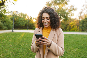 Wall Mural - Young african happy woman walking outdoors in a spring park using mobile phone.