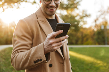 Sticker - Photo of smiling caucasian man listening to music with earpods and typing on smartphone in autumn park