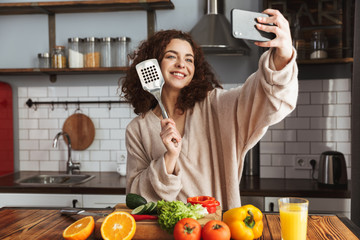 Image of joyful caucasian woman taking selfie photo on smartphone while cooking at home