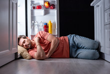Exhausted Man Sleeping On Floor In Kitchen Near Open Refrigerator