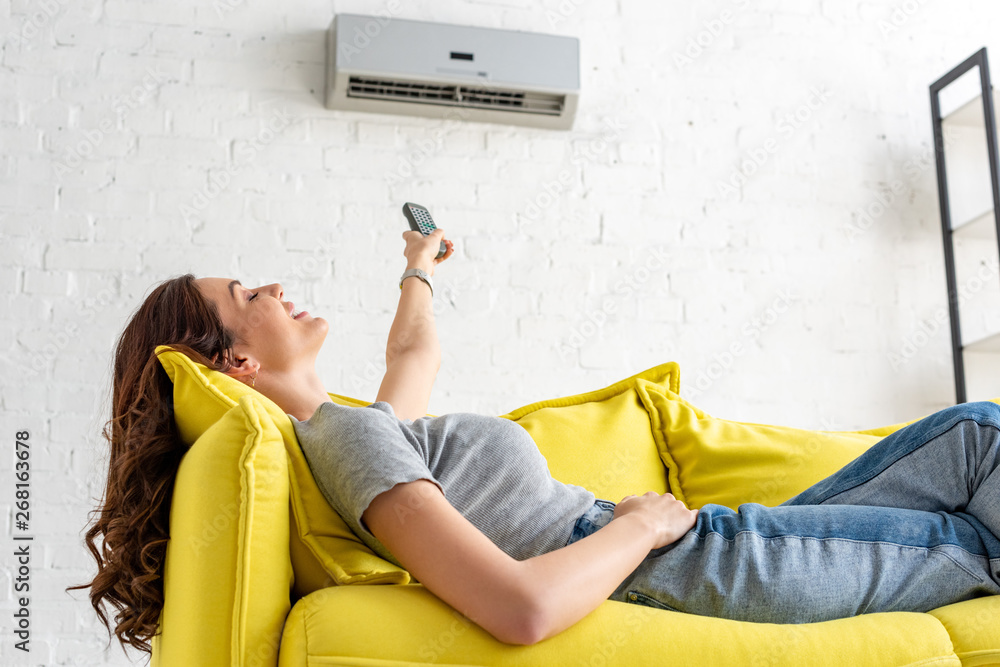 attractive young woman relaxing under air conditioner and holding remote control - obrazy, fototapety, plakaty 