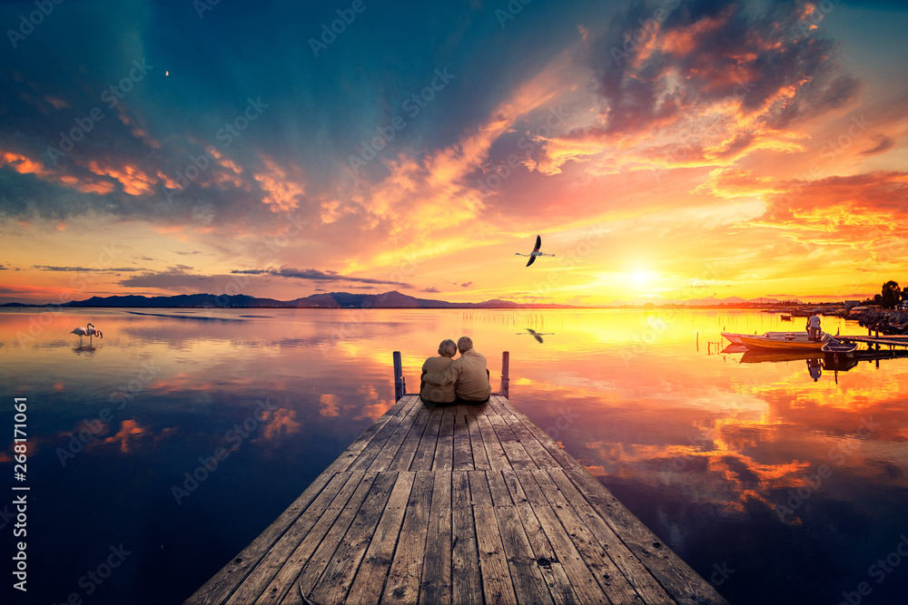 Senior couple seated on a wooden jetty, looking a colorful sunset on the sea with a flying flamingo reflected on the calm water. - obrazy, fototapety, plakaty 