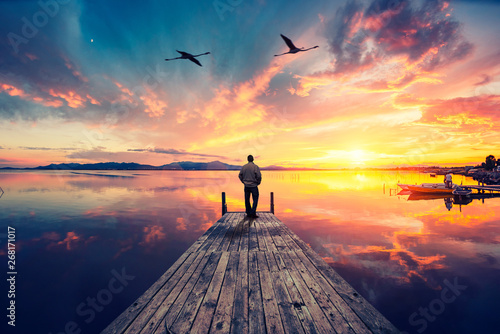 Cuadros en Lienzo Lonely man on wooden pier contemplating a colorful sunset reflected on the sea with two flamingos flying high