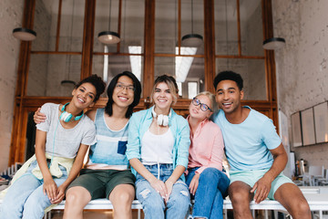 Wall Mural - Smiling asian young man in glasses gently embracing girl with light-brown skin. Indoor portrait of glad students having fun in library while preparing for exams.