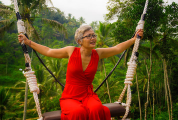 Wall Mural - natural lifestyle portrait of attractive happy middle aged 40s - 50s Asian woman with grey hair and stylish red dress riding rainforest swing carefree swinging