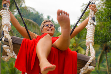 Wall Mural - attractive happy middle aged 40s or 50s Asian Indonesian woman with grey hair riding rainforest swing carefree swinging and enjoying tropical jungle adventure