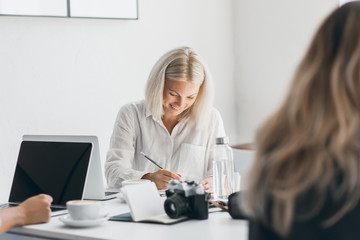 Wall Mural - Laughing blonde woman in white shirt looking down while writing something. Indoor portrait of busy female freelance specialist posing at workplace with laptop and camera.