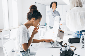 Wall Mural - Pensive black girl sitting in earphones near laptop while her colleagues standing beside flipchart. Indoor portrait of female photographer working with computer, typing on keyboard in office.